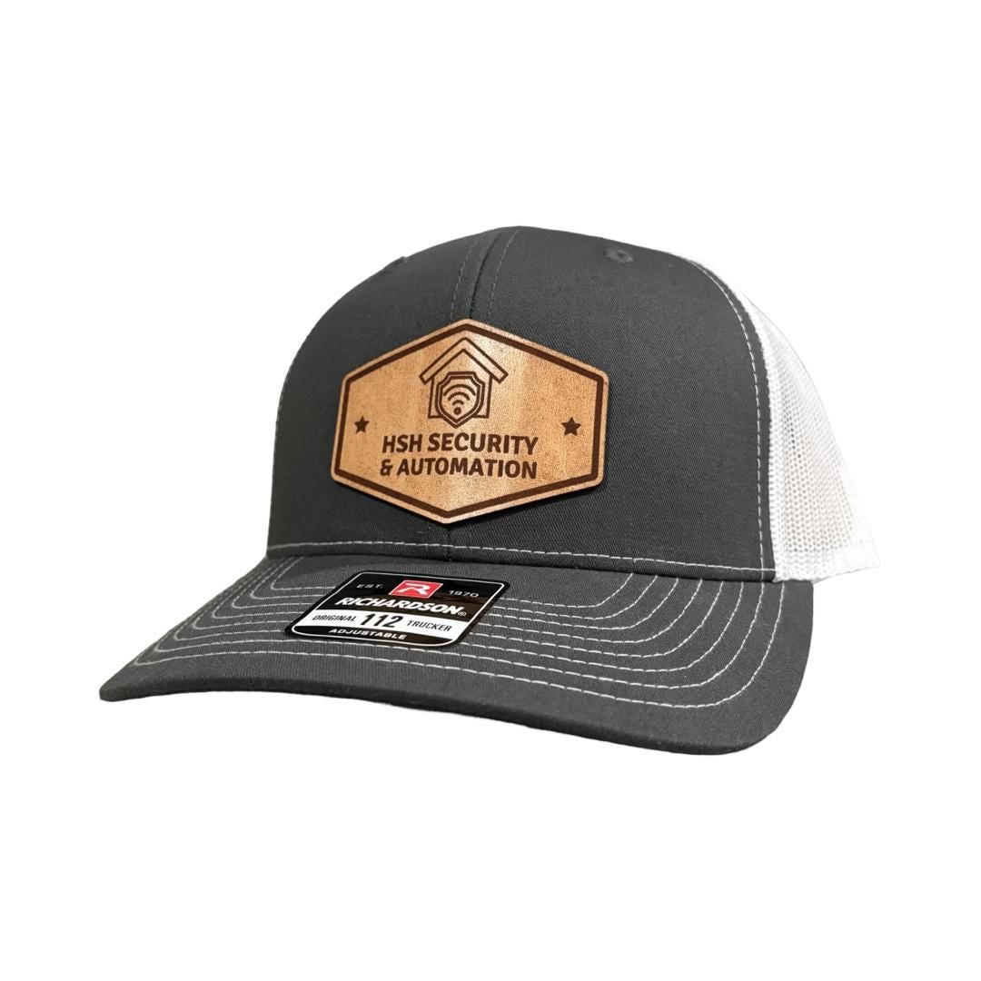 A Charcoal Gray and White Richardson 112 Trucker hat with a hexagonal leather patch showcasing a security company