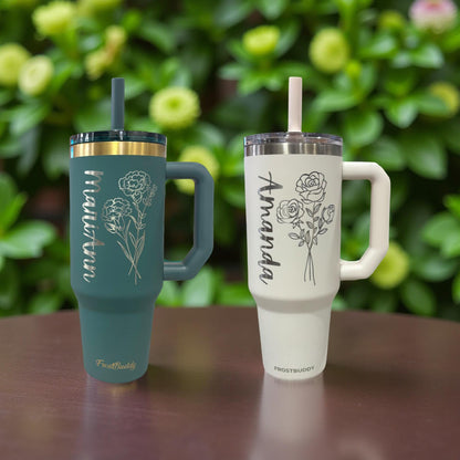Two insulated Personalized and Laser Engraved Frost Buddy Tumblers with Birth Flower designs and names on a wooden surface with a greenery and flowers in the background.